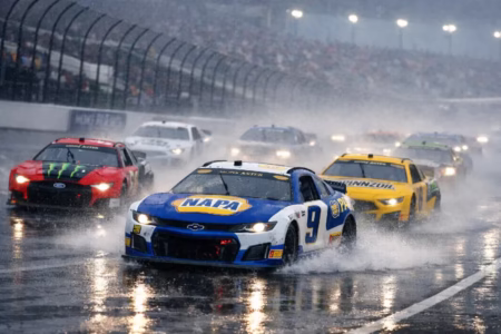 NASCAR stock cars racing at high speed on a rain‑soaked track, headlights cutting through mist as water spray rises behind them.